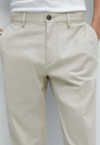 Pantaloni beige in cotone con texture liscia. Presentano una chiusura con bottone e tasche laterali. Design su misura con una vestibilità dritta.