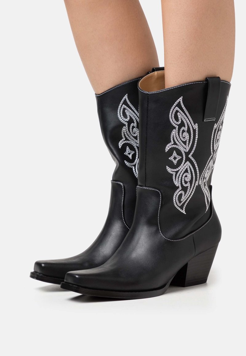 Bottes de cowboy en cuir noir mi-mollet avec des coutures décoratives blanches et des talons empilés, portées sur des jambes nues, sur fond blanc.