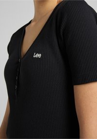 Sort ribbet top med dyb V-hals, korte ærmer og fem-knaps lukning. Har broderet "Lee" logo på brystet.