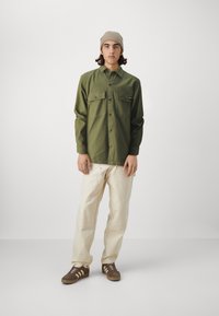 Green button-up shirt with two front pockets, cream trousers, and brown sneakers. Model wears a light knit beanie, standing against a neutral background.