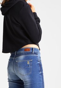 Kratší černá mikina s kapucí a šňůrkou a modré džíny s koženou cedulkou a jemným vyblednutím na zadních kapsách.