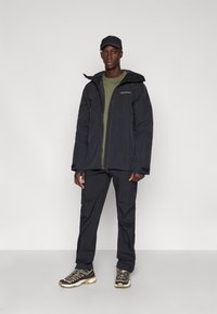 Casaco preto impermeável com capuz, camisa verde, calças pretas e sapatos de caminhada com um design texturizado e detalhes em contraste.