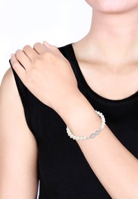 Armband aus weißen Perlen mit einem silbernen Unendlichkeitssymbol, am Handgelenk getragen. Der Hintergrund ist schlicht weiß, was das Schmuckstück hervorhebt.