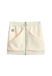 Minifalda beige con cremallera plateada en el frente, cinturilla elástica, bolsillos laterales inclinados y un pequeño parche redondo blanco y negro en el lado izquierdo.