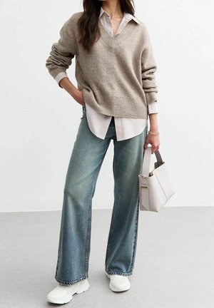 Femme portant un pull beige par-dessus une chemise rayée, jean large, baskets blanches, tenant un sac à main crème, debout devant un fond uni.
