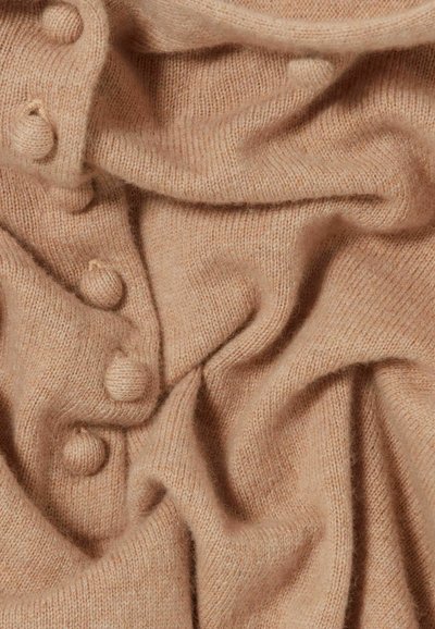 Tissu en tricot beige clair avec une texture douce, présentant des boutons ronds et un drapé délicat, créant une apparence cocooning et superposée.