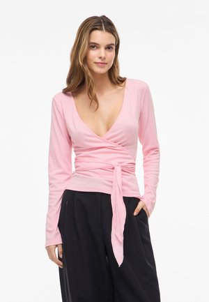 Γυναίκα που φοράει ένα ανοιχτό ροζ τοπ τύπου wrap με μακριά μανίκια και ζώνη δεμένη στη μέση, συνδυασμένο με φαρδιά μαύρη παντελόνια, στέκεται με το ένα χέρι στην τσέπη.