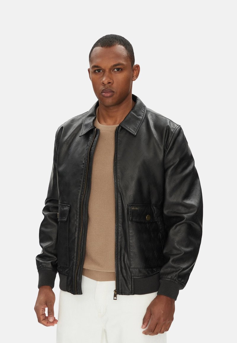 Giacca bomber in pelle nera con una texture liscia, chiusura con zip, due tasche frontali e polsini a coste. Indossata sopra un top beige.