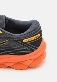 Scarpa sportiva nera e grigia con suola arancione. Presenta una tomaia in mesh testurizzato, dettagli gialli e un design ondulato sui lati.