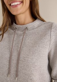 Sweatshirt gris clair avec un col montant, doté d'un tissu texturé, de cordons de serrage et d'œillets métalliques pour un ajustement réglable.