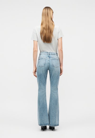 Γυναίκα με μακριά ξανθά μαλλιά, που φοράει ανοιχτό γκρι μπλουζάκι, ψηλόμεσα ανοιχτό μπλε τζιν με καμπάνα και μαύρες ψηλοτάκουνες γόβες, στέκεται με την πλάτη της προς τα εμπρός.