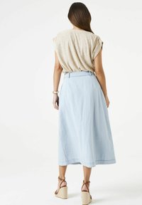 Hellblauer Jeans-Maxi-Rock mit taillierter Taille und dezenter Stichung, kombiniert mit einem beigen kurzärmeligen Oberteil und braunen Keilsandaletten.