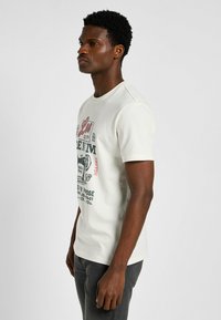 Camiseta de algodón crema con mangas cortas, cuello redondo y estampado gráfico frontal en rojo y verde. Combinada con jeans gris oscuro.