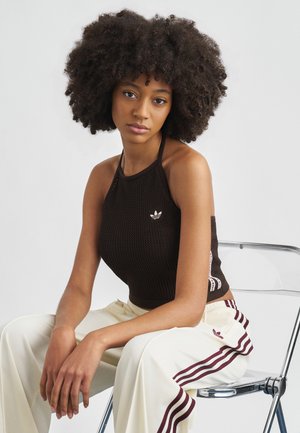 Καφέ τοπ-χιαστί από υφασμένο πλεκτό ύφασμα με μικρό λευκό λογότυπο. Το μοντέλο φοράει κρεμ παντελόνι με φαρδιά μπατζάκια και μπορντό ρίγες. Φόντο με διαφανή καρέκλα.