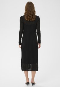 Robe noire en tricot côtelé avec de longues manches, longueur mi-mollet et texture transparente. Coupe droite sans fermetures visibles. Détail fente à l'ourlet.