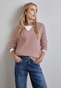 Ribbad rosa tröja med V-ringning, lager på lager över en vit skjorta. Modellen bär blå jeans, med händerna i fickorna och står något vänd åt vänster.