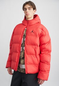 Joven vestido con una chaqueta acolchada roja brillante con capucha, parcialmente abrochada sobre una camiseta gráfica, de pie contra un fondo liso claro.