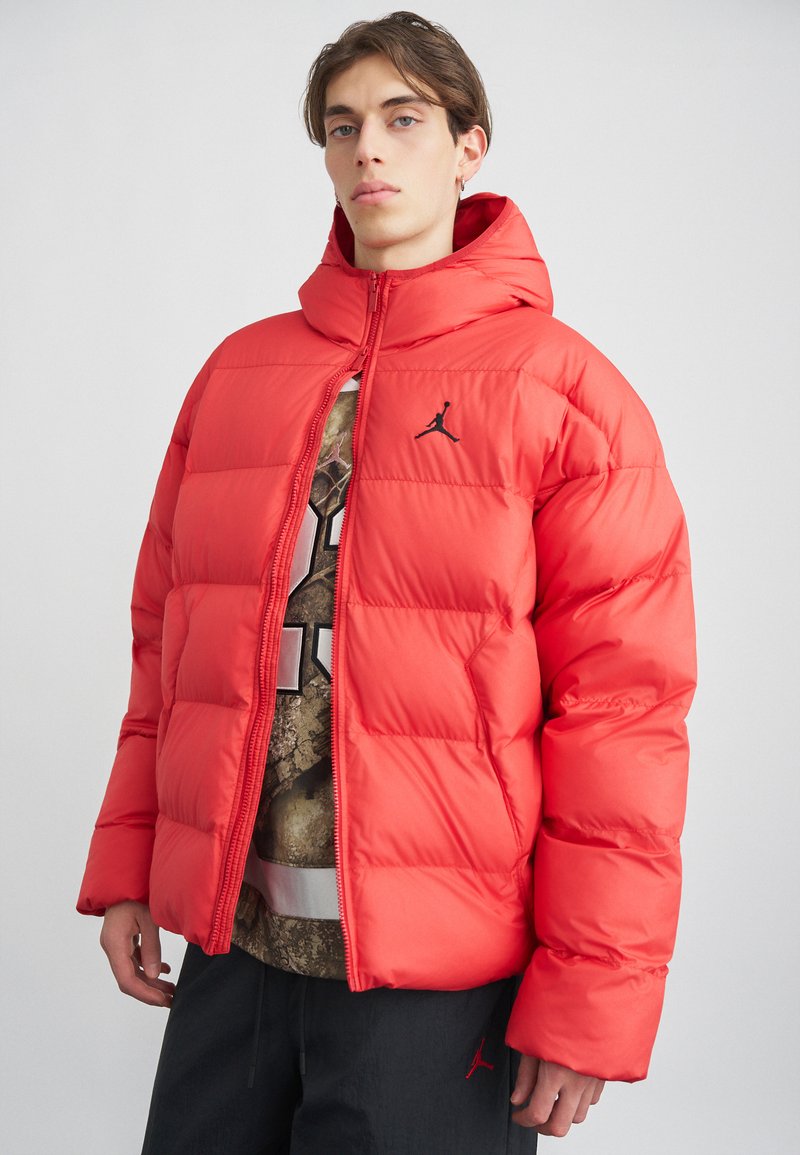 Joven vestido con una chaqueta acolchada roja brillante con capucha, parcialmente abrochada sobre una camiseta gráfica, de pie contra un fondo liso claro.