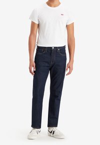 Mörkblå denimjeans med rak benpassform, femfickorsdesign och kontrasterande orange sömmar, tillsammans med en vit T-shirt med kort ärm.