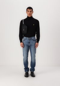 Pulover negru cu guler înalt, blugi albaștri și pantofi negri. Logo mic galben pe pulover. Geantă crossbody cu curea cu text alb.