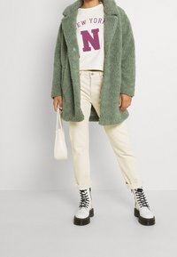 Persona che indossa un cappotto verde texturizzato, un maglione crema del New York College, pantaloni beige chiari, stivali bianchi lacciati, mentre tiene una piccola borsa bianca.