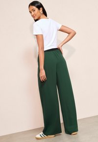 Olijfgroene wijde broek met een zwarte elastische tailleband, gecombineerd met een witte T-shirt met korte mouwen. De schoenen hebben een crème basis met groene strepen.