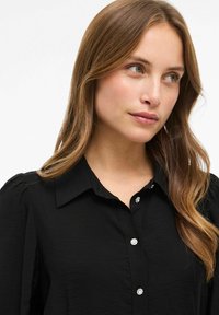 Crna košulja s dugim rukavima i ovratnikom, izrađena od teksturirane tkanine, s napuhanim rukavima i dekorativnim kristalnim dugmadi duž kopčanja.