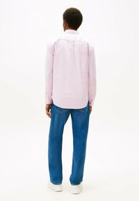 Camicia rosa chiaro a maniche lunghe con dettaglio di bottoni, indossata con jeans blu e sneaker bianche. La camicia ha un orlo arrotondato ed è leggermente oversize.