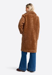 Brauner, flauschiger, übergroßer Mantel mit Kragen und langen Ärmeln, kombiniert mit blauen Schlagjeans und braunen Schuhen, von hinten betrachtet.
