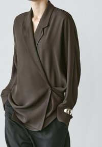 Person trägt eine lockere, dunkelbraune Wickelbluse mit einem Knopf, schwarze Hose und ein silbernes Armband am linken Handgelenk.