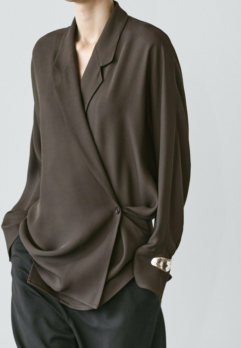 Person trägt eine lockere, dunkelbraune Wickelbluse mit einem Knopf, schwarze Hose und ein silbernes Armband am linken Handgelenk.