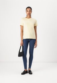 Lys gul bomull t-skjorte med korte ermer, med hvit tekstlogo. Kombinert med blå skinny jeans og svarte loafers. Holder en svart håndveske.