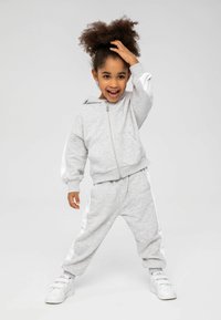 Ensemble de survêtement de sport gris avec des bandes latérales blanches, comprenant un hoodie zippé et un pantalon décontracté. Le modèle porte des sneakers blanches.
