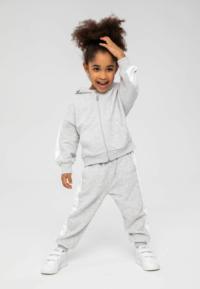 Ensemble de survêtement de sport gris avec des bandes latérales blanches, comprenant un hoodie zippé et un pantalon décontracté. Le modèle porte des sneakers blanches.