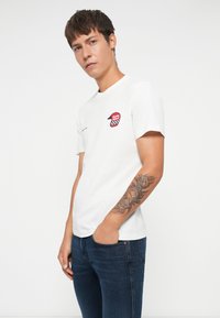 Camiseta de algodón blanca con un parche gráfico a cuadros en rojo y negro, escote redondo, mangas cortas, combinada con jeans azul oscuro.