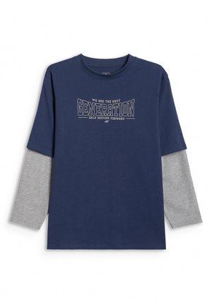 Chłopiec w granatowym, za dużym T-shircie na szarym długim rękawie, szarych spodniach dresowych i szarych butach sportowych z różowymi sznurówkami, stoi na jednolitym tle.