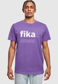 Fialové bavlněné tričko s krátkým ruk�ávem, na kterém je velkým bílým písmem napsáno slovo "fika" a pod tím je popisný text. Volný střih.