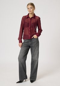 Sheer burgundy lace button-up shirt with a diamond pattern, featuring long sleeves and a collared neckline, paired with gray wide-leg jeans.