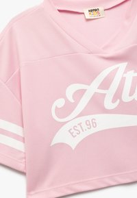 Rosa, kortærmet trøje lavet af mesh-stof. Har hvide striber på ærmerne og et stort hvidt tekstdesign, der siger "At" med "EST.96" nedenfor.