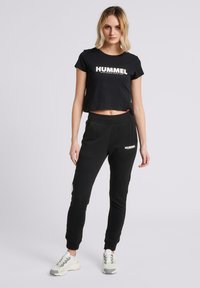 Camiseta negra corta con el logo "HUMMEL", combinada con joggers negros que presentan detalles en blanco y una cintura elástica, finalizados con zapatillas blancas.