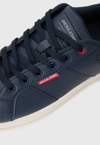 Navy blue sneaker with perforated synthetic upper, round laces, red logo tag, and white rubber sole. Features a cushioned collar for comfort.