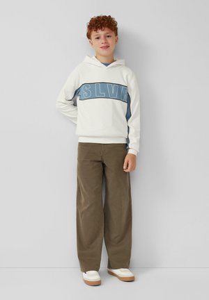 Un garçon aux cheveux bouclés portant un hoodie blanc avec un imprimé bleu "SLVR", un pantalon large marron et des baskets blanches, se tenant devant un mur uni.