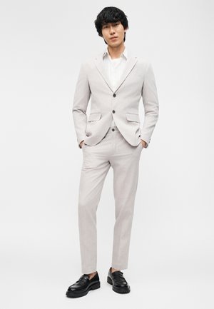 Man standing with hands in pockets wearing a light gray suit, white shirt, and black loafers against a plain light background.