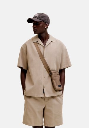 Hombre con camisa beige de manga corta abotonada, pantalones cortos a juego, bolso cruzado y gorra de béisbol oscura sobre un fondo claro y sencillo.
