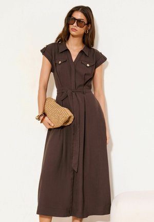 Nő sötétbarna, öves midi ruhát visel, amelynek a mellrészen gombos zsebei vannak, fonott kézitáskát tart a kezében, napszemüveget és arany karika fülbevalókat visel.