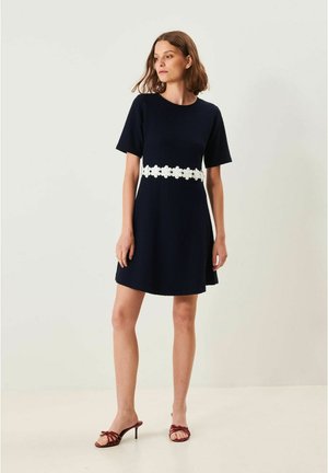 Kvinna som bär en marinblå klänning med kort ärm och vitt blommönster vid midjan samt röda öppna klackskor, ståendes mot en enfärgad bakgrund.