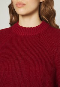 Femme portant un pull en maille côtelée rouge avec un col rond, visible du menton jusqu'au haut de la poitrine.
