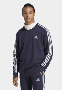 Tamno plavi Adidas dukserica s bijelim trostrukim trakama na rukavima i logom na prsima. Tkanina izgleda meko s opuštenim krojem.