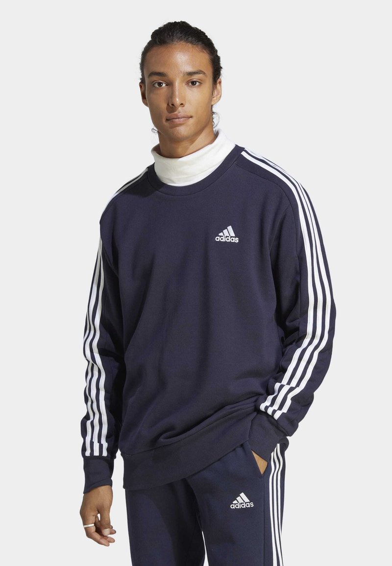 Tamno plavi Adidas dukserica s bijelim trostrukim trakama na rukavima i logom na prsima. Tkanina izgleda meko s opuštenim krojem.