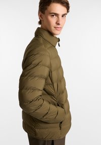 Chaqueta acolchada verde oliva con cuello alto, patrón acolchado, bolsillos con cremallera y un tejido ligero y texturizado.